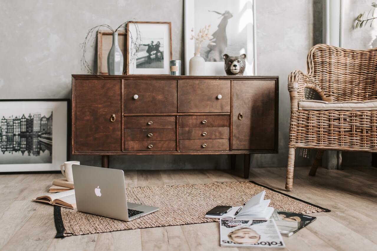 Une pièce dans une maison avec une armoire de style simpliste en bois légèrement usé, un tapis avec un ordinateur portable, des magazines et des livres sur le sol, des peintures ornant les murs et une chaise en osier.